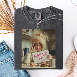 Op de afbeelding: Donkergrijze Comfort Colors t-shirt met een afbeelding van een pop met een bord met de tekst "Nailed It". Een zilveren ketting en witte schoenen zijn ook zichtbaar. Het shirt is opgevouwen op een wit oppervlak met een spijkerbroek en een gedroogde palmblad.