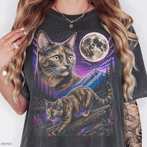 Pode incluir: Uma t-shirt cinza-carvão com uma estampa gráfica de dois gatos tartaruga num cenário noturno. O design inclui uma lua cheia grande, nuvens roxas e azuis e uma cordilheira. Um gato é mostrado em retrato, o outro a caminhar sobre um tronco de árvore.