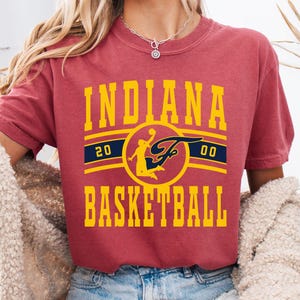Puede incluir: Una camiseta granate con un gráfico amarillo y azul marino que dice "INDIANA BASKETBALL". El gráfico incluye una silueta de jugador de baloncesto y los números "20" y "00".