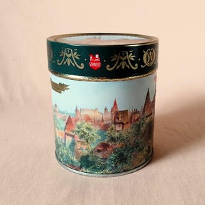 Könnte beinhalten: Eine zylindrische Dose mit einer detaillierten Landschaftsszene mit Gebäuden und Bäumen. Die Dose hat einen dunkelgrünen Deckel mit goldenen Akzenten und dem Wort "Schmidt" in Rot. Die Farbpalette umfasst Blau-, Grün-, Braun- und Goldtöne.