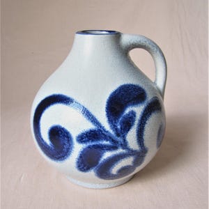 Könnte beinhalten: Weißer Keramikkrug mit rundem Körper und Henkel. Der Krug ist mit einem blauen Blumenmuster verziert. Der Krug hat eine kleine Öffnung oben.