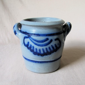 Könnte beinhalten: Ein kleiner, hellblauer Keramiktopf mit zwei Henkeln. Der Topf hat ein dunkelblaues Dekorationsmuster in der Mitte und ein dunkelblaues Band in der Nähe des oberen und unteren Randes. Der Topf wird wahrscheinlich zu Dekorationszwecken verwendet.