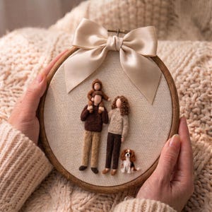 May include: A circular wooden embroidery hoop containing a needle-felted family portrait on light fabric. The scene features two adults, a child sitting on an adult's shoulders, and a small tricolor dog, all topped with a large beige satin bow.