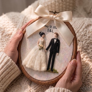 Può includere: Un telaio da ricamo rotondo in legno contenente figure di sposi in lana infeltrita su tessuto bianco. La sposa indossa un abito bianco e lo sposo un abito nero. Sopra c'è un fiocco in raso color crema e il testo May. 12. 2024 S&L.