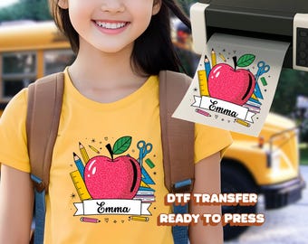 May include: A yellow t-shirt featuring a large pink apple design with the name "Emma" on a banner. The apple is surrounded by school supplies like pencils, scissors, and a ruler. The words "DTF TRANSFER READY TO PRESS" are also visible.