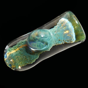 May include: A clear glass pipe with a rounded bowl and a textured, colorful interior. The interior features shades of teal, green, and gold, creating a unique, artistic design. The pipe is set against a black background.