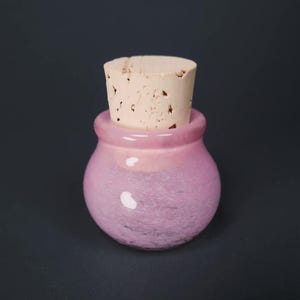 May include: A small, round, pink glass jar with a cork stopper. The jar has a bulbous body and a short neck. The cork is light brown and fits snugly in the jar's opening. The jar is a soft pink color with a slightly textured appearance.