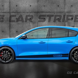 May include: A vibrant blue Ford Focus hatchback, featuring black racing stripes and black alloy wheels. The car displays the word "FOCUS" on its side. The background is a grey concrete wall with the text "OZ CAR STRIPES".
