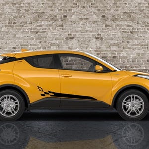 May include: A yellow Toyota C-HR compact SUV with black racing stripes and a checkered flag design on the side. The car has silver alloy wheels and is parked in front of a brick wall.