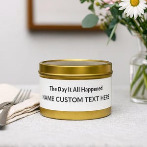以下が含まれることがあります： 白いラベルが付いた金色のキャンドル缶。「The Day It All Happened」と「NAME CUSTOM TEXT HERE」の文字が黒で書かれています。キャンドルは、フォークと折りたたまれたナプキンが置かれた灰色の表面にあります。