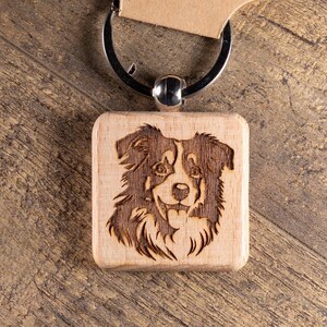 May include: A wooden keychain featuring a laser-etched image of a dog's face. The dog has brown and white fur. The keychain is attached to a metal ring and a brown cardboard tag with the text "T&R Creative Concepts LLC".