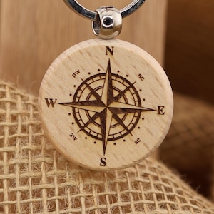 May include: A wooden compass keychain with a silver keyring. The compass rose is etched into the light-colored wood. The keychain hangs from a brown tag with the text "T&A Creative Concepts LLC QUALITY PRODUCTS" and the website address.