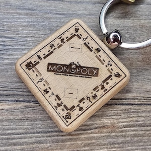 May include: A wooden Monopoly game board keychain with a silver key ring and a brass key. The keychain is square-shaped with the Monopoly logo and game board details etched into the wood. The key is attached to the ring.