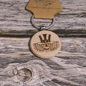 May include: A wooden keychain with a round pendant featuring an engraved skull wearing a top hat. The keychain includes a metal ring and a cardboard tag. The website uscreativeconcepts.com is printed on the tag.