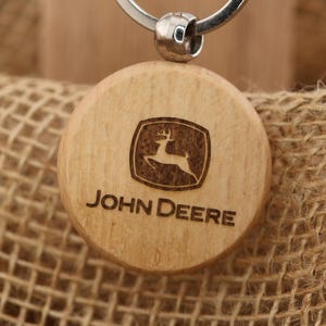 May include: A wooden John Deere keychain with a silver keyring. The keychain features the John Deere logo and text engraved on a round wooden disc. The keychain is attached to a brown cardboard tag with the T&A Creative Concepts LLC logo and website.