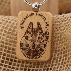 Pode incluir: Porta-chaves de madeira com uma gravura detalhada da Millennium Falcon e as palavras "Millennium Falcon Keys". O porta-chaves inclui um anel prateado e uma etiqueta com o texto "T&A Creative Concepts LLC Quality Products" e um endereço web.