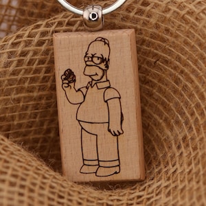 Może przedstawiać: Drewniany brelok do kluczy z postacią z kreskówki trzymającą pączka. Brelok ma srebrny pierścień i brązową zawieszkę z napisem "T&A Creative Concepts LLC QUALITY PRODUCTS" oraz stroną internetową tacreativeconceptsllc.com.