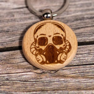 May include: A wooden keychain with a laser-etched design of a skull wearing a gas mask. The keychain has a silver metal ring and a brown cardboard tag. The skull design is in a dark brown color.