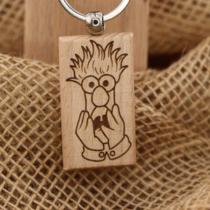 May include: Wooden keyring with a cartoon character engraved on a rectangular block. The keyring has a silver ring and a brown tag with the text "T&A Creative Concepts LLC QUALITY PRODUCTS" and the website address.