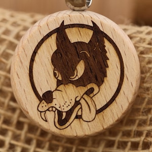 May include: A round wooden pendant with a cartoon wolf design. The wolf is etched in dark brown within a circular frame on the light-colored wood. The wolf has its tongue out. A silver metal loop is at the top.