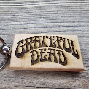 May include: A wooden keychain featuring the words "Grateful Dead" in a stylized font, burned into the wood. The keychain has a silver metal ring and ball. The wood has a light, natural color.
