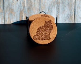 May include: Set of four round cork coasters with a black metal holder. Each coaster features a detailed, etched design of a cat with swirling patterns. The coasters are a natural brown color.