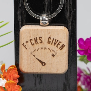 May include: A wooden keychain with a silver ring. The square wooden pendant has the words "F*CKS GIVEN" engraved, along with a gauge graphic. The gauge reads 100. The keychain is attached to a black wooden backing.