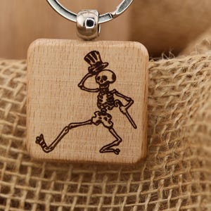 Puede incluir: Llavero de madera con un esqueleto grabado con láser que lleva un sombrero de copa y sostiene un bastón. El esqueleto está representado en una pose de caminar. El llavero tiene un anillo de metal plateado.