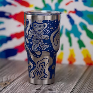 May include: A stainless steel tumbler with a blue and silver design. The tumbler features a stylized bear graphic with the words "Grateful Dead" and "Love" incorporated into the design. The background is a tie-dye pattern.