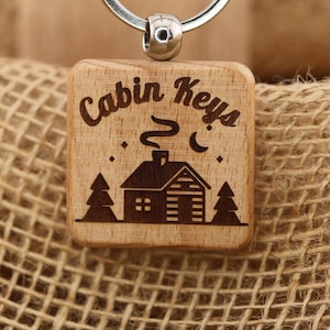 May include: Wooden keyring with a square design. The keyring features the words "Cabin Keys" above an etched image of a cabin, two trees, a crescent moon, and stars. The keyring is attached to a silver metal ring.