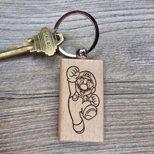 Può includere: Un portachiavi in legno con un'immagine incisa al laser di Mario del videogioco Super Mario Bros. Il portachiavi è rettangolare e attaccato a un portachiavi argentato. È presente anche una chiave in ottone.