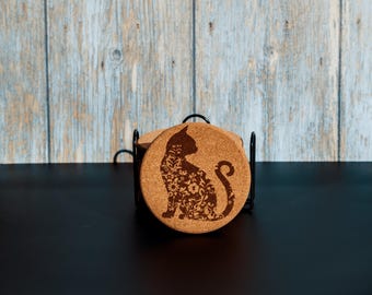 May include: Set of round cork coasters with a black metal holder. Each coaster features a detailed silhouette of a cat filled with floral patterns. The coasters are a natural cork color, and the holder is black.