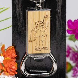 May include: A silver bottle opener keyring with a rectangular bamboo insert featuring a cartoon character. The character is holding a bottle. The keyring is attached to a black wooden background with flowers.