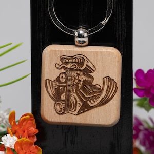 Puede incluir: Llavero cuadrado de madera con una imagen grabada de un motor de hot rod. El llavero tiene un anillo de metal plateado y está sujeto a una pantalla de madera negra. El fondo incluye flores.