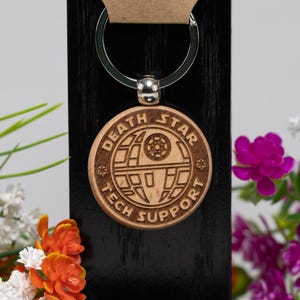 May include: A wooden Death Star keychain with the words "DEATH STAR TECH SUPPORT" engraved on it. The keychain is attached to a silver keyring and a small tag with the website www.tacreativeconceptsllc.com.