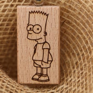 Może przedstawiać: Prostokątna drewniana zawieszka z grawerowanym laserowo wizerunkiem Barta Simpsona z serialu Simpsonowie. Zawieszka jest jasnobrązowa i wisi na sznurku, na tle juty.