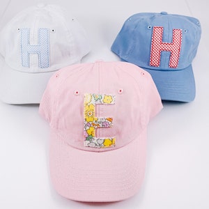 May include: Three baseball caps in white, light blue, and pink. Each cap features a large, embroidered letter: "H" in blue gingham on the white cap, "H" in red gingham on the blue cap, and "E" in a floral pattern on the pink cap.