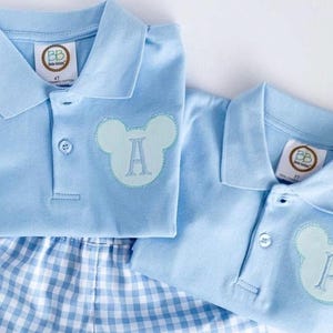May include: Two light blue polo shirts with a Mickey Mouse-shaped applique, one with the letter "A" and the other with the letter "P". The shirts are paired with blue and white checkered shorts. Each shirt has a tag that says "Beba Beliza".
