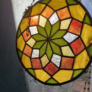 Puede incluir: Una vidriera redonda con un diseño geométrico. La ventana presenta una estrella verde central, rodeada de piezas de vidrio amarillas, naranjas, rojas y blancas. Una cadena de metal está unida para colgarla.