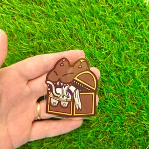 May include: A clear sticker featuring a cartoon alligator emerging from a treasure chest. The alligator is brown with white teeth and a white scarf. The treasure chest is brown with a gold trim.