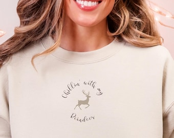 May include: A cream-colored sweatshirt with the text "Chillin' with my Reindeer" and a reindeer graphic. The person is wearing light blue jeans and a reindeer-themed slipper. A white and red mug with a gingerbread man is also visible.