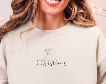 May include: A cream-colored sweatshirt with the text "merry Christmas" in a delicate script. The sweatshirt features a small floral design above the text. A red and white mug with a gingerbread man design is held in the hand. The person is wearing light blue jeans and a reindeer slipper.