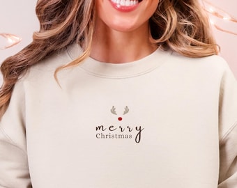 May include: A cream-colored sweatshirt with the text "merry Christmas" in a script font, with a small reindeer design above the text. The person is wearing light blue jeans and a pair of reindeer slippers. A white mug with a gingerbread man is held in the person's hand.
