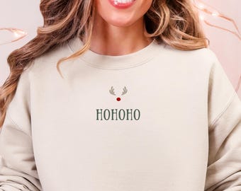 May include: A beige sweatshirt with a small reindeer design and the word "HOHOHO" in green. The person is wearing light blue jeans and a pair of Christmas-themed slippers. A white mug with a gingerbread man is held in the person's hand.