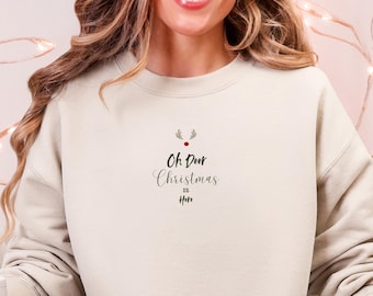 May include: A cream-colored sweatshirt with the text "Oh Deer Christmas is Here" in black script. The sweatshirt has a small reindeer graphic above the text. The person is wearing light blue jeans and a pair of reindeer slippers.