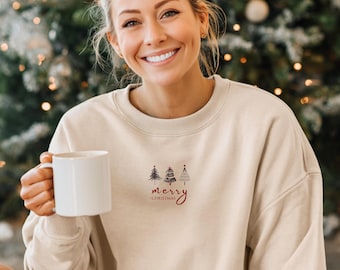 May include: Beige sweatshirt with a small graphic of three Christmas trees and the words "merry christmas" in red script. The sweatshirt is worn by a person holding a white mug.