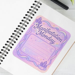 Może przedstawiać: Spiralny notatnik z naklejką "Manifestation Monday" w odcieniach fioletu i różu, ozdobioną kryształami i księżycami. Widoczny jest również czarny aparat fotograficzny i długopis.