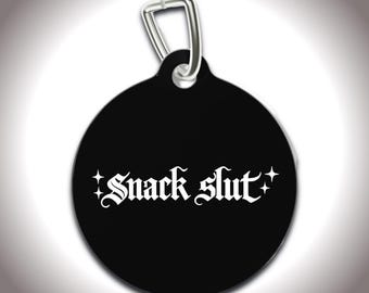May include: Black, round pet tag with a silver clasp. The tag features the white text "Snack slut" in a gothic font, with star accents on each side of the text.