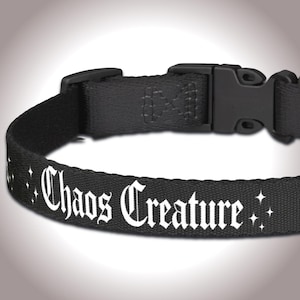 Pode incluir: Coleira preta para animais de estimação com o texto "Chaos Creature" em fonte gótica branca. A coleira tem uma fivela de plástico preta e é decorada com pequenas estrelas brancas.