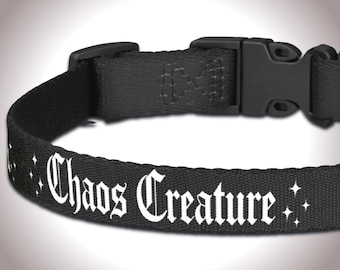 May include: Black pet collar with the text "Chaos Creature" in white gothic font. The collar has a black plastic buckle and is decorated with small white starbursts.